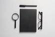 Notebook with black leather cover, eyeglasses, pen and magnifying glass on gray background. Flat lay