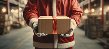 Santa Claus In A Shipping Warehouse With A Cardboard Box In His Hands - Concept Of Online Shopping And Home Deliveries At Christmas
