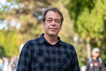 Wall Mural - Street portrait of a relaxed 40-50 year old man wearing a shirt against a neutral blurred background of a city park. Perhaps he is a shopper, actor or lorry driver, military pensioner.