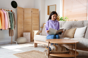 Sticker - Beautiful young Asian woman in stylish hoodie sitting on sofa and reading magazine at home