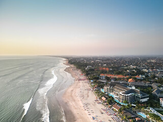 Aerial view of Beautiful sunset on the double six beach with Hotel, resort, cafe buildings, beach umbrella and calm ocean wave in Bali. Tourists are surfing, swimming and walking along the beach.