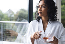 Happy Biracial Woman In Bathrobe Holding Cup Of Tea And Looking Out Window In Sunny Room At Home
