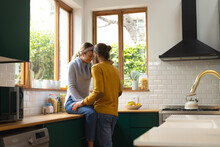 Happy, Romantic Caucasian Couple Embracing Sitting On Worktop And Standing In Kitchen, Copy Space