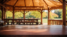 A Wooden Gazebo With A Thatched Roof In A Park. The Gazebo Has A Wooden Table And Benches. In The Background Is A Blurred View Of The Park With Trees And Grass