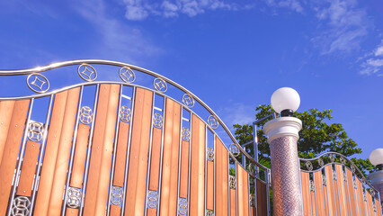 Wall Mural - Low angle view of modern stainless steel and wooden sliding gate door with round lamp on mosaic pillar in front of home against blue sky background