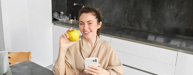 Wall Mural - Close up portrait of smiling brunette woman in bathrobe, sits in kitchen at home, uses mobile phone and holds an apple, orders fresh fruits on smartphone app, buys groceries online, looks up recipe