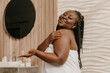 © gstockstudio - Happy plus size African woman covered in towel applying cosmetic cream on shoulder in bathroom