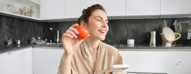 Wall Mural - Young woman thinks what to cook, sits in the kitchen with smartphone and tomato in hands, looks aside and smiles, searches recipes on mobile app, orders groceries