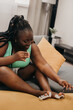 © gstockstudio - Beautiful plus size African woman painting toenails and smiling while sitting on the couch at home