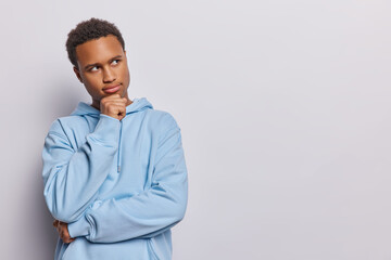 Wall Mural - Horizontal shot of handsome curly haired African man keeps hand under chin concentrated aside pensively thinks over offer wears casual blue sweatshirt poses over white background. Let me think concept