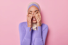 Horizontal Shot Of Positive Muslim Woman Keeps Eyes Closed Keeps Hands On Face Smiles Gladfully Wears Traditional Hijab Laughs At Something Funny Isolated On Pink Background. Positive Emotions Concept