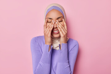 Wall Mural - Horizontal shot of positive Muslim woman keeps eyes closed keeps hands on face smiles gladfully wears traditional hijab laughs at something funny isolated on pink background. Positive emotions concept