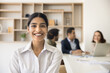 © fizkes - Happy young beautiful Indian businesswoman office portrait. Positive pretty business professional girl looking at camera, posing in co-working meeting room with colleagues talking behind