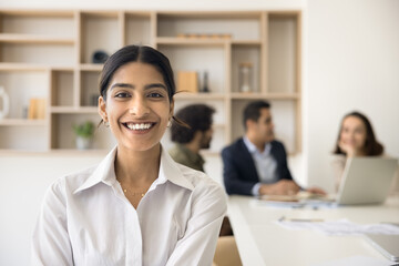 Wall Mural - Happy young beautiful Indian businesswoman office portrait. Positive pretty business professional girl looking at camera, posing in co-working meeting room with colleagues talking behind