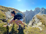 Fototapeta Las - Sporty woman on a trek in the Dolomites, black shirt, shorts, dark hair, backpack, sunny day. Physically demanding climb, rocks, mountains peaks in the background.