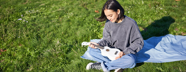 Smiling asian girl, musician playing ukulele in park, looking with care and love at her instrument, sitting on blanket on sunny day