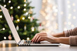 a person typing on a laptop in front of a christmas tree with white lights behind it and a lit green christmas tree