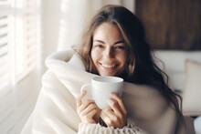 Young woman wrapped in a white blanket  holding a cup with hot drink trying to warm up in the cold apartment