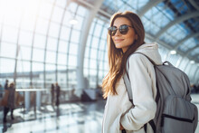 Young Woman At The Airport Wearing A Light Jacket, Sunglasses And A Backpack.