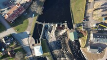 Aerial View Of A Big Dam, Barrage, Weir In A City Near A Green And Residential Area On A Sunny Day. Water Flowing.