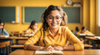 Smiling young female student wearing yellow shirt and eyeglasses, sitting in class with her hands laid on a notebook, school board and other students blurred in the background