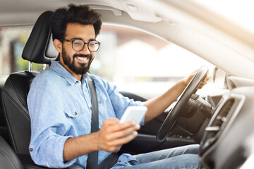 Wall Mural - Cheerful young indian guy driver using smartphone while driving car