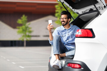 Wall Mural - Cheerful indian guy sitting in open car trunk, using smartphone