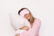 © Марина Демешко - A young caucasian sleepy smiling blonde woman in a pink pajamas and sleep mask on her eyes hugging a pillow isolated on a white background. Insomnia, sweet dreams, cuddling with a pillow