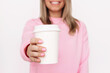 © Марина Демешко - Cropped shot of a young blonde smiling woman in a pink sweatshirt holding out her hand with white eco paper cup with tea or coffee with copy space for text or design isolated on a light background