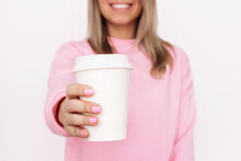 Cropped Shot Of A Young Blonde Smiling Woman In A Pink Sweatshirt Holding Out Her Hand With White Eco Paper Cup With Tea Or Coffee With Copy Space For Text Or Design Isolated On A Light Background