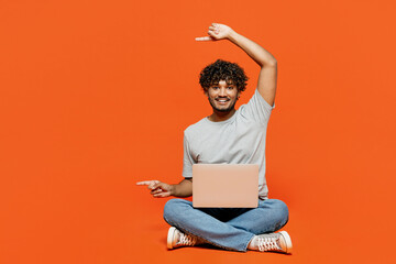 Full body young happy IT Indian man he wear t-shirt casual clothes sit hold use work on laptop pc computer point finger aside isolated on orange red color background studio portrait Lifestyle concept