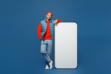 Sticker - Full body young smiling happy man in padded vest red hat stand near big huge blank screen mobile cell phone smartphone with area looking camera isolated on plain dark blue background studio portrait