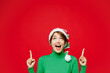 © ViDi Studio - Young woman wear green turtleneck Santa hat posing point index finger overhead indicate on workspace area isolated on plain red background. Happy New Year 2024 celebration Christmas holiday concept.