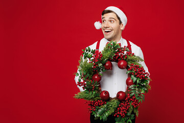 Wall Mural - Merry smiling young man wear white shirt Santa hat posing hold Christmas wreath look aside on area mock up isolated on plain red background studio portrait. Happy New Year celebration holiday concept.