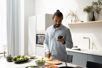 Serious young Black chef guy reading recipe on mobile phone, consulting food blog, website, cooking dinner from organic food ingredients, cutting vegetables for salad, making order with online app