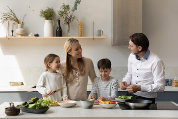 Cheerful couple of parents and two sibling kids cooking fresh food together, cutting vegetables for salad at kitchen table, talking, laughing, sharing household chores. Family healthy eating concept