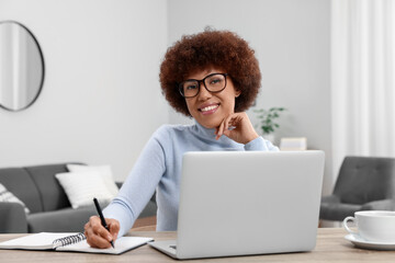 Poster - Beautiful young woman using laptop and writing in notebook at wooden desk in room