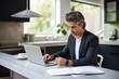 © radekcho - Smiling elegant businessman holding documents and looking at camera while working from home. Freelancer working from home sitting at desk with laptop. Self employed business person working from home.