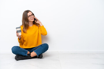 Wall Mural - Young caucasian woman sitting on the floor isolated on white background holding coffee to take away and a mobile