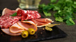 Fresh slices cold cuts, sausage ham bacon jamon on stone cutting board with olives on background of greenery. Meat pork beef. Food