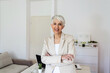 © Jelena Stanojkovic - Confident female looking at camera while standing in home office. Cropped portrait of an attractive senior businesswoman sitting at the desk with her arms crossed while working from home.