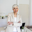 © Jelena Stanojkovic - Confident female looking at camera while standing in home office. Cropped portrait of an attractive senior businesswoman sitting at the desk with her arms crossed while working from home.