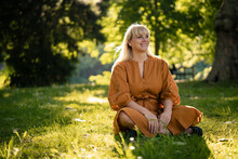 Happy Blond Woman Sitting On Grass In Park