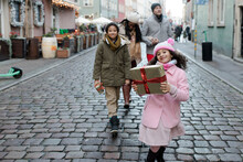 Happy Girl Holding Gift Box And Walking With Family At Christmas Market