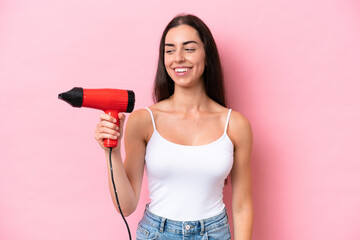 Wall Mural - Young caucasian woman holding a hairdryer isolated on pink background with happy expression