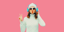 Winter portrait of stylish modern woman in wireless headphones listening to music wearing white knitted sweater and hat, sunglasses, posing on pink studio background