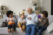 © fizkes - Great-grandpa having fun with three little great-grandchildren, sit together on sofa and play musical instruments, celebrate family holiday, loving grandsons congratulate older relative with birthday