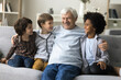© fizkes - Three little multiethnic brothers spend time together with mature great-grandpa, hugging, resting on sofa enjoy talk and harmonic relationships at home. Multigenerational family ties, bond, attachment