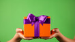 Anonymous person showing bright orange carton gift box with ribbon and purple bow against green background