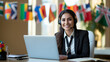 Beautiful smiling multilingual female interpreter wearing a headset with microphone, sitting at her desk with a laptop, international flags behind her